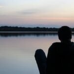 Adult sitting by water at sunset contemplating life choices and childfree regret.
